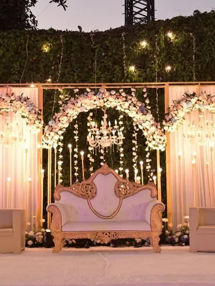 A sophisticated reception stage with a royal touch. The ornate white and gold sofa is the centerpiece, set against a backdrop of floral arches and hanging crystal strings.