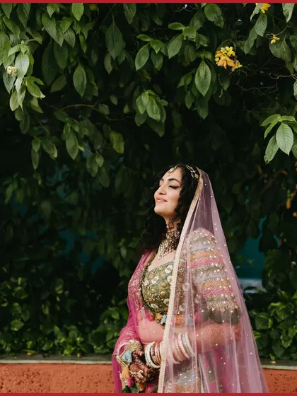 A serene portrait of a bride with her eyes closed, basking in the sunlight, her pink veil softly framing her face against a green backdrop.