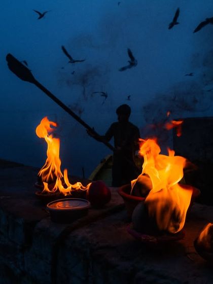 A boatman paddles on the Yamuna river at dawn, silhouetted against the flames of a ritual fire on the ghat.
