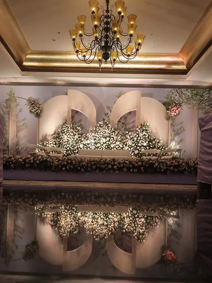 This stage reflects modern artistry with its sculptural white panels and a mountain of lush, pastel florals. The glossy, reflective floor doubles the visual impact, creating a dreamy and sophisticated atmosphere for a wedding reception.
