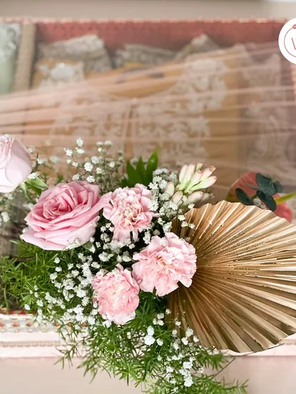 A bespoke trousseau packaging box in a lovely shade of pink. The clear lid is adorned with a delicate arrangement of pink roses, carnations, and a golden palm leaf for a touch of modern elegance.