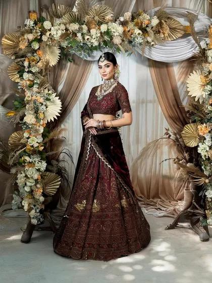 A full-length shot of a bride in a stunning maroon velvet lehenga. The overall look is one of grandeur, with the makeup designed to complement the rich fabric and elaborate floral decor.