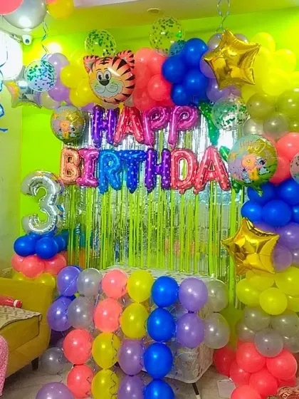 A colorful home birthday decoration for a 3-year-old, with a bright green fringe curtain and a multi-colored balloon setup.