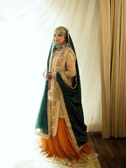 A full-length portrait of a Muslim bride in her stunning orange and green attire. The natural light from the window highlights her elegance and the richness of her outfit.