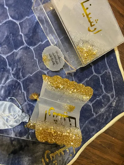 A personalized gift set featuring a dome-shaped prayer mat in a beautiful blue pattern. The set is completed with a custom resin Quran box and holder, a crystal tasbeeh, a bookmark, and a car hanging, all in a coordinated white and gold glitter design.