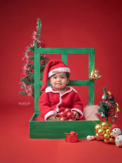 A cozy Christmas portrait with a baby dressed as Santa sitting inside a green window frame prop. The solid red background and festive ornaments create a classic holiday card look.