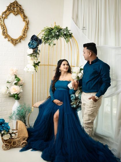 A regal portrait of an expecting couple. The mother-to-be is seated in the golden birdcage chair, wearing a magnificent royal blue gown, creating a striking and elegant image.