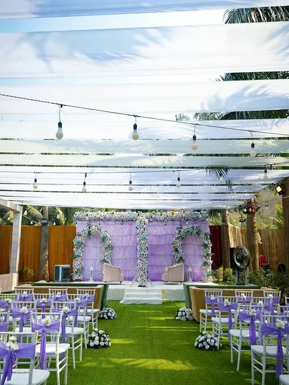 A wide shot of the lavender-themed wedding stage. The floral arches against the draped backdrop and the neat rows of guest seating create a serene and organized look.