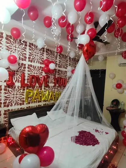 A classic romantic surprise that never fails. This bedroom decoration includes red and white balloons covering the ceiling, 'I LOVE YOU' foil letters, a delicate canopy over the bed, and a heart made of rose petals.