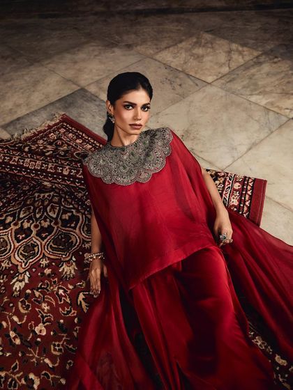 A model lies gracefully on a rug, draped in a stunning red cape set with a heavily embellished silver collar.