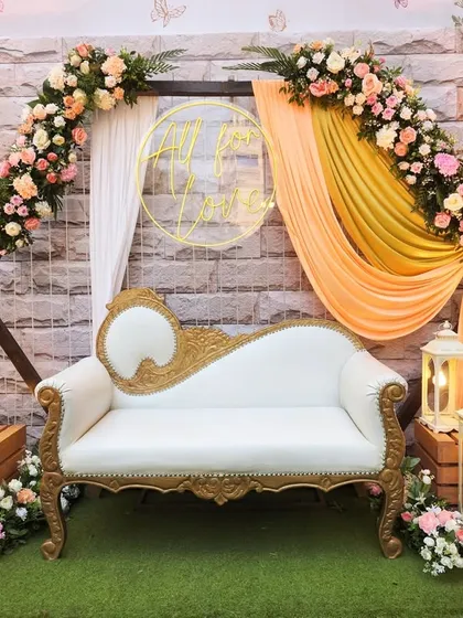 This photo booth combines a floral-printed backdrop with a soft, draped arch and a custom "All for Love" neon sign. The classic white and gold diwan completes this romantic setup.
