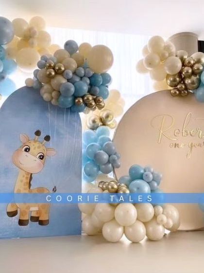 A sweet and gentle theme for a first birthday celebration. This setup features a cute giraffe illustration on a custom arch, surrounded by a soft blue, white, and gold balloon arrangement for a memorable milestone.