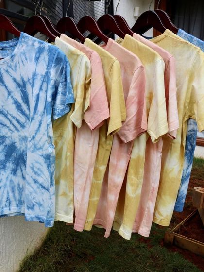 A rack of my naturally dyed t-shirts, available for purchase. Each one is hand-dyed with natural pigments like indigo, marigold, and sappanwood, resulting in unique patterns and colours.