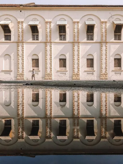 A creative composition showing a building and its perfect reflection in a puddle. A person walking by is also reflected, creating a symmetrical and surreal image that plays with perception.