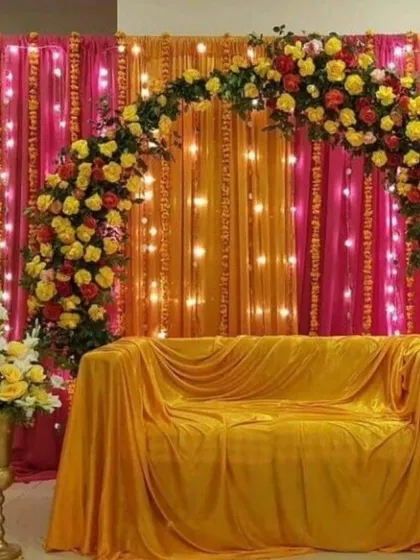 A beautiful photo corner for a Haldi or Mehandi event, featuring a floral arch with yellow and red roses over a sofa covered in yellow fabric. The backdrop of pink and orange drapes with fairy lights adds a warm glow.
