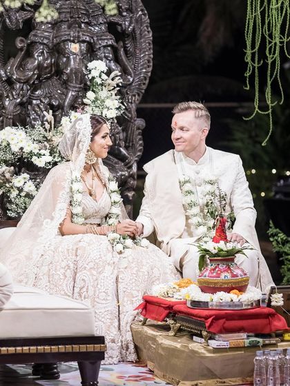 A beautiful moment captured at the mandap, where the couple sits before a magnificent Ganesha sculpture. The decor features a canopy of white flowers and hanging floral strings, creating a serene and blessed atmosphere for their wedding vows.