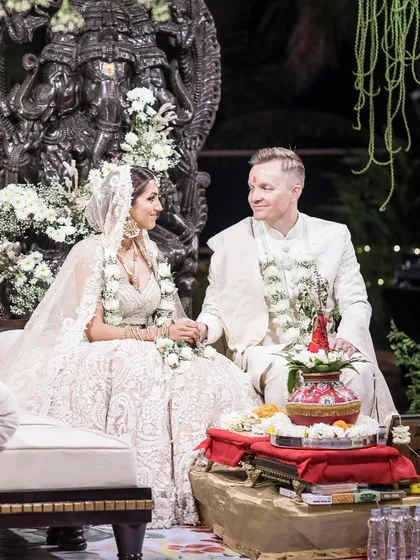 A beautiful moment captured at the mandap, where the couple sits before a magnificent Ganesha sculpture. The decor features a canopy of white flowers and hanging floral strings, creating a serene and blessed atmosphere for their wedding vows.