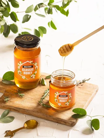 A bright, clean shot for Melfons honey, showing the product's pure, viscous texture as it drips from a honey dipper. The use of fresh green leaves in the background adds a natural, organic feel.