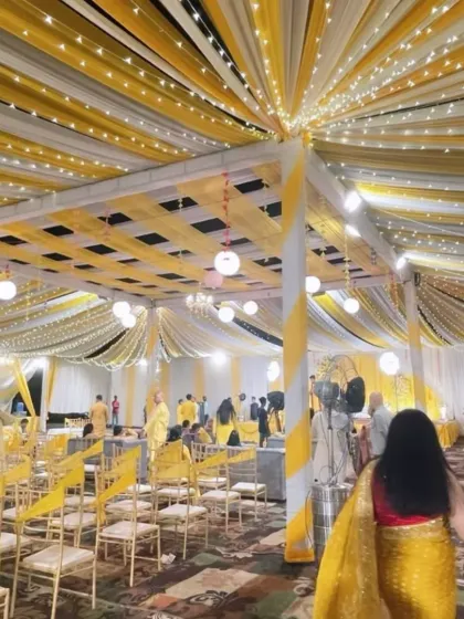An indoor Haldi event setup with a yellow and white theme. The ceiling drapes are lit with fairy lights, and the guest seating is arranged to face the main stage, ensuring everyone is part of the celebration.