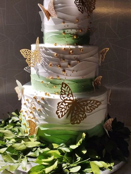 A stunning four-tiered cake with a green ombre effect and delicate gold butterflies. The base is surrounded by fresh green leaves, creating a beautiful natural aesthetic.