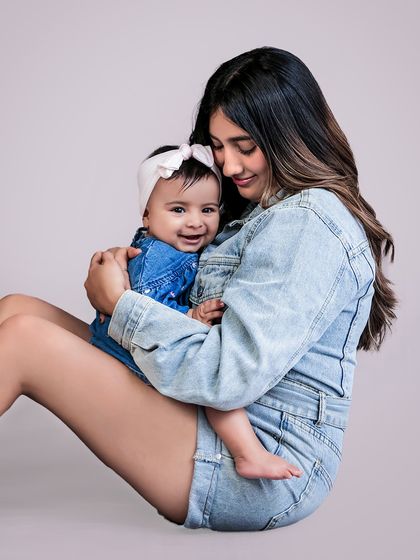 A mother holds her baby close, both sharing a beautiful smile. The coordinated denim outfits create a classic, cohesive look for this parent-child portrait.