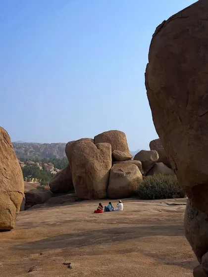 A few people sit and relax on a vast expanse of rock, dwarfed by the giant boulders around them. The image conveys the immense scale and peacefulness of the Hampi landscape.