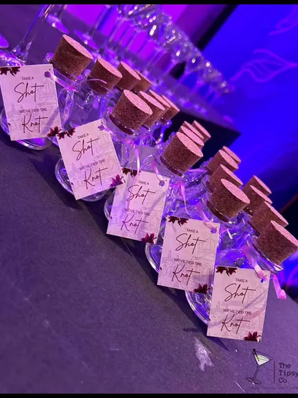 A close-up of our custom shot bottles with personalized tags that say 'Take a Shot, We Tied The Knot'. The purple lighting adds to the party atmosphere.