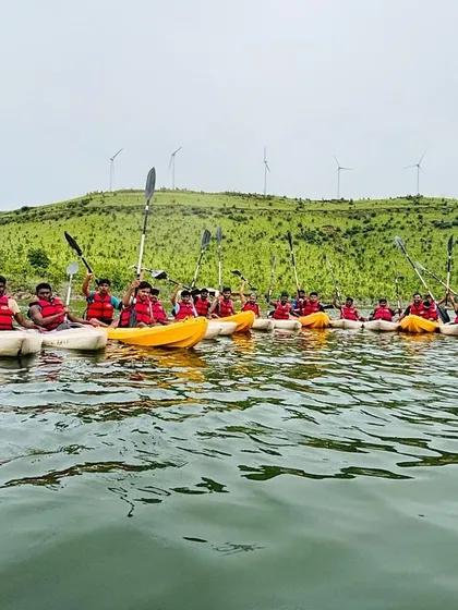A large group of kayakers raises their paddles for a photo, with the iconic windmills of the Chitradurga region in the background.