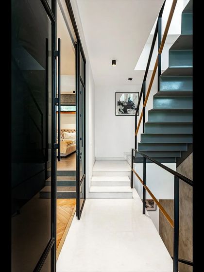 A narrow corridor in 'SOL' leading towards a bedroom, with the steel staircase visible alongside. The clean lines and minimalist material palette of white walls, dark steel, and glass doors create a serene and modern feel.