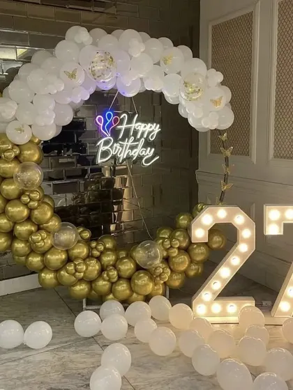 A chic 27th birthday setup featuring a semi-arch of gold and white balloons against a mirrored wall, paired with large marquee numbers.