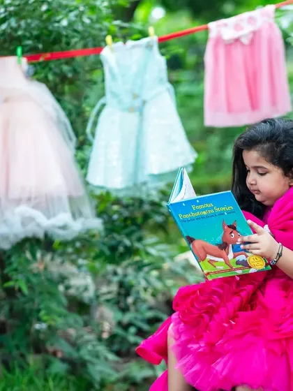 A whimsical outdoor photo of a young girl in a bright pink dress, engrossed in a storybook with little dresses hanging on a line behind her.