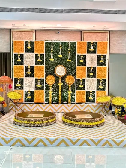 The main seating area for a Haldi, featuring a backdrop of checkered marigold panels and a chevron-patterned floor. The low traditional seating (bajots) completes the look.