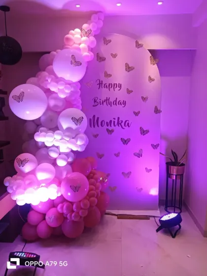 A lovely pink and purple butterfly-themed decoration. An arched backdrop is adorned with butterfly stickers and a cascading balloon garland, all illuminated by soft purple lighting.