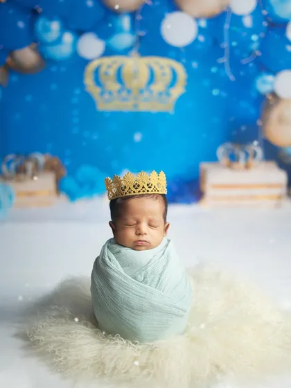 A beautifully composed shot of the newborn prince, swaddled and crowned, against a backdrop of blue and gold balloons.