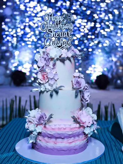 The 25th-anniversary cake in its element, set against a sparkling blue backdrop at the celebration. The silver "Happy 25th Anniversary" topper adds the final touch.
