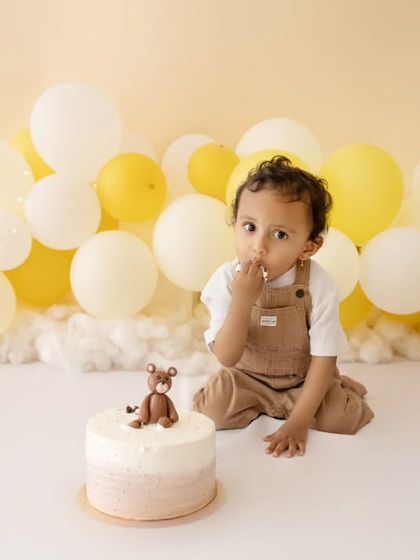 The moment before the smash. A sweet portrait of a baby boy with his teddy bear cake, set against a backdrop of yellow and white balloons.