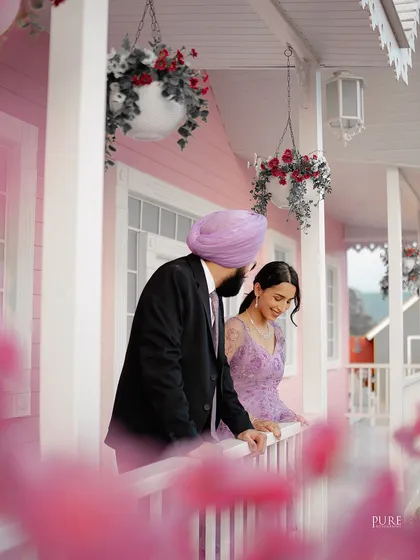 A beautifully soft and romantic shot of Manpreet and Simranpreet on the porch of a pastel pink house set. The foreground flowers add depth and a dreamy quality to the photo.