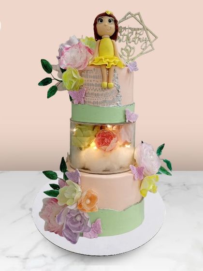 A beautiful three-tiered cake with a separator tier filled with flowers and lights, topped with a sweet doll figure.