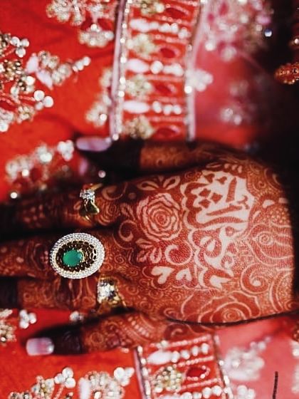 A close-up shot from a professional wedding photoshoot, highlighting the intricate details of the stained mehendi against the bride's red lehenga.