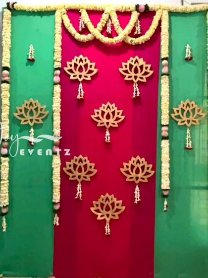 A simple yet elegant backdrop for a half saree function, featuring wooden lotus cutouts on a dual-tone pink and green fabric. The look is completed with hanging jasmine garlands.