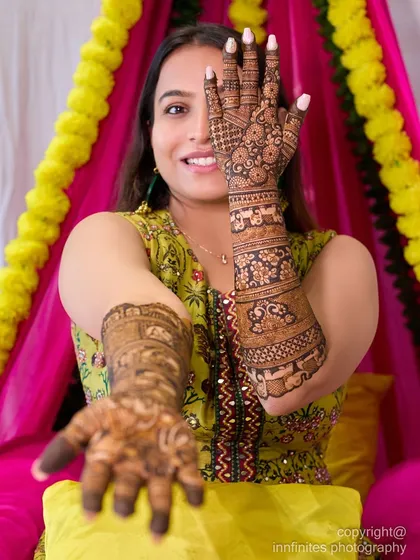 A classic bridal portrait for a Mehndi ceremony, where the bride playfully shows off her intricate henna designs. The shot is framed to highlight both her joyful expression and the detailed artistry on her hands and arms.
