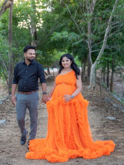 A happy couple walking hand-in-hand through a wooded path. The mom-to-be's bright orange gown adds a beautiful pop of color to the natural scenery.