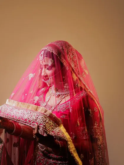 This portrait captures the bride in a moment of quiet reflection, looking down at her veil. The warm, soft lighting enhances the rich color of her red lehenga and the intricate embroidery.