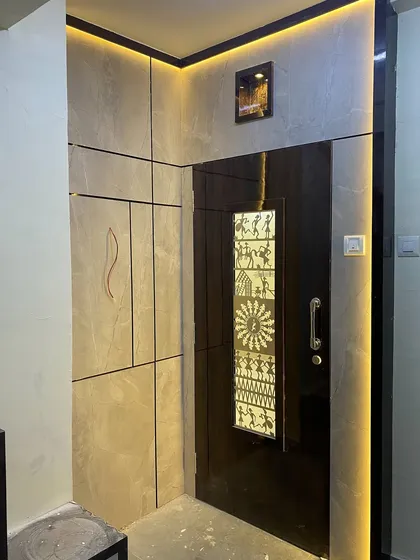 A sophisticated entrance with a dark door featuring a backlit Warli art panel. The surrounding grey wall panels and strip lighting create a dramatic and modern look.