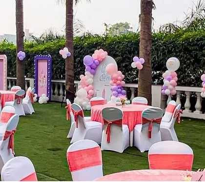 An outdoor princess party setup with pink and white table settings. The background features themed backdrops and pastel balloon decorations, transforming the lawn into a fairytale kingdom.