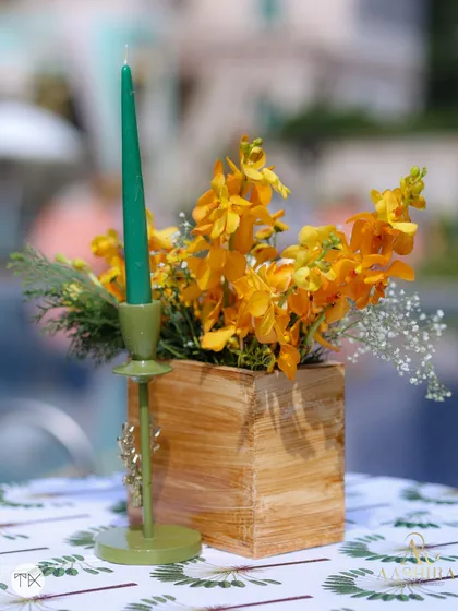 A simple yet elegant detail of a wooden box with yellow orchids, paired with a green candle, on a leaf-printed tablecloth.