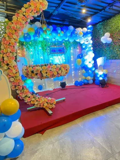 A beautiful naming ceremony decoration with a cradle filled with flowers, set on a stage with a green wall and blue and yellow balloons.