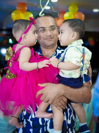A father holds his two young children, capturing a candid interaction between the siblings at a party.