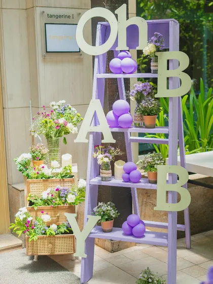 This 'Oh Baby' ladder shelf is a charming and versatile decor piece. Dressed in a soft lavender hue and adorned with small floral pots and balloon accents, it serves as a perfect welcome element or a creative photo prop for your baby shower.