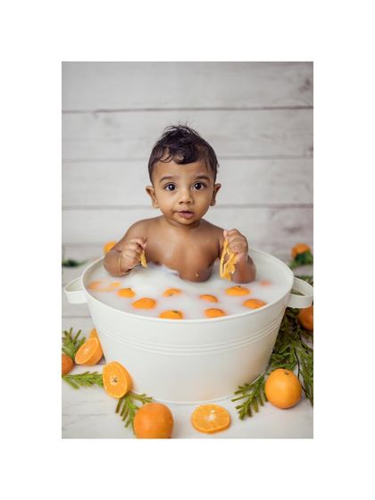 A baby boy in a milk bath plays with orange slices. His direct gaze at the camera creates an engaging and adorable portrait.